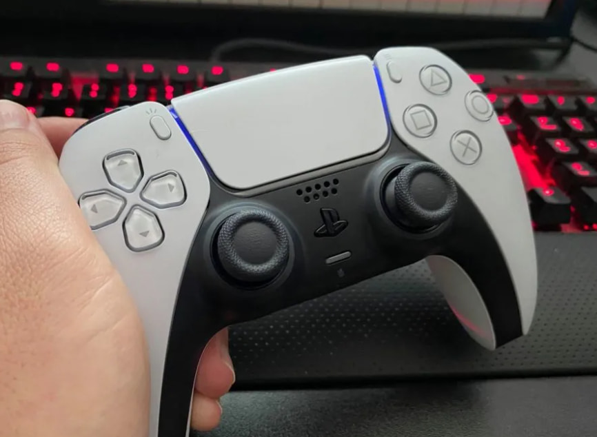 A PS5 controller in a person's hand against a red-lit RGB keyboard.