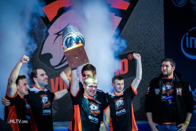 NEO from Virtus.pro lifting the EMS One Katowice trophy on stage with his teammates lifting their hands in the air.