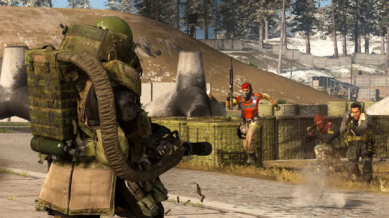 A juggernaut soldier is aiming a minigun at soldiers that are pursuing him. The soldiers are armed with guns, while there are trees and crates behind them. 