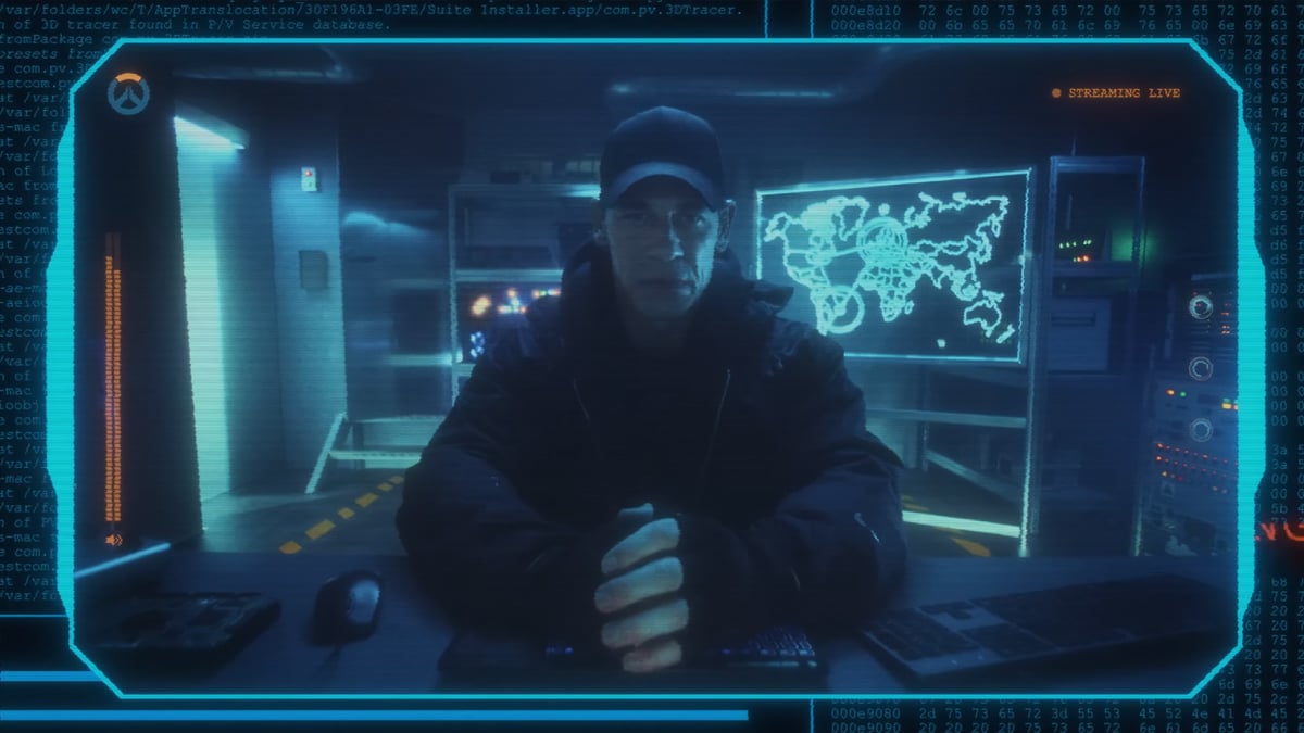 Film and TV star John Cena in a hoodie and cap, sitting in a room and hacking a computer for Overwatch 2.