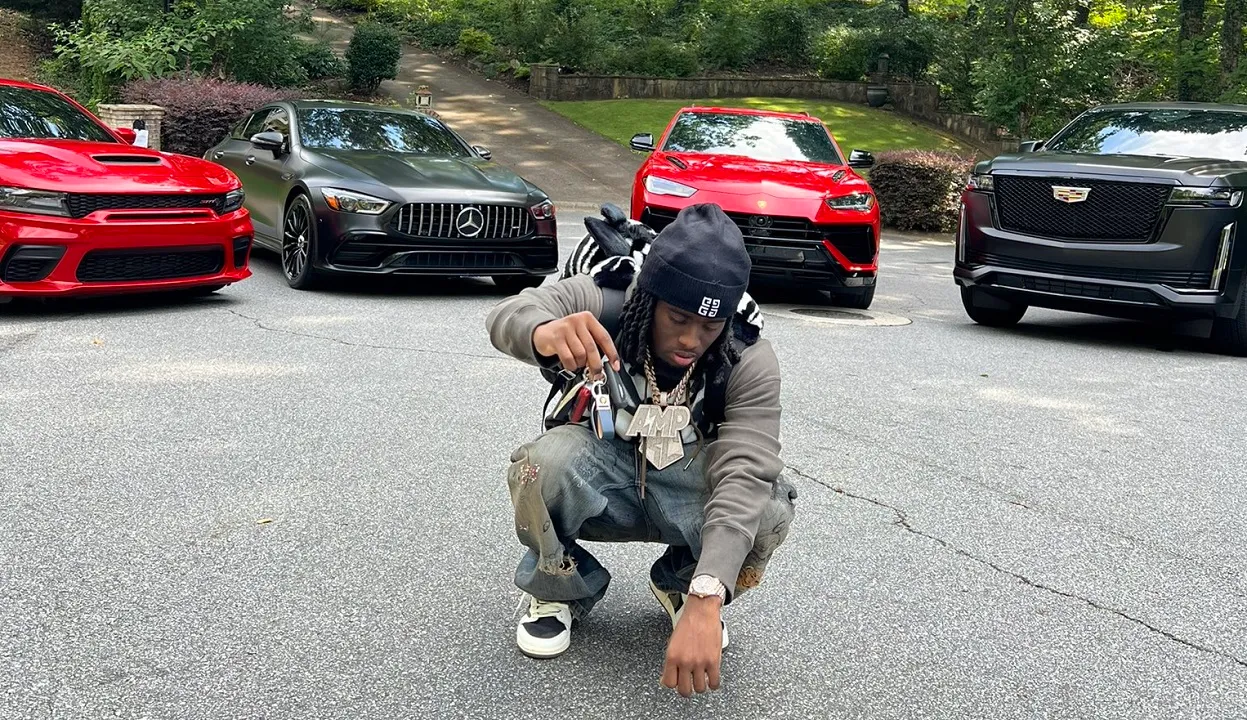 There is a picture of Kai Cenat sitting in front of multiple cars he owns. There are two red cars and two black cars behind him. 