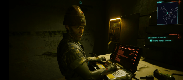 V speaks to a contact for a mission wearing a beanie and on a computer in Cyberpunk 2077: Phantom Liberty. 