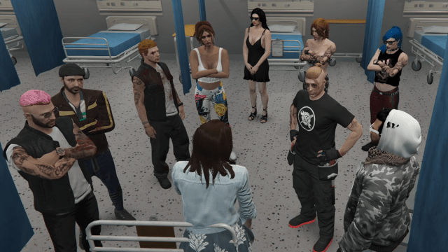 A group of characters in GTA 5 stand in a room in the hospital, all looking at a woman leaning on the bed.