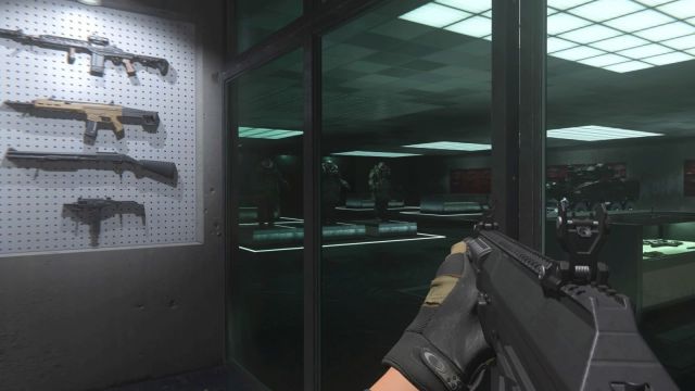 Image showcasing the firing range backroom in MW3 (2023). There is a weapon on show in the frame with others held on a side wall. Beyond tinted glass there are juggernaut armours standing with a vehicle nearby.