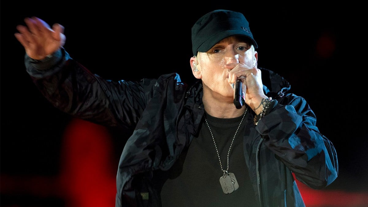 Eminem wearing a black cap, jumper, and jacket, holding a microphone rapping.