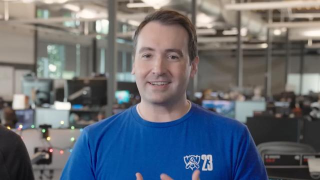 A man with short, brunette hair and a blue t-shirt stands in front of a busy office space.