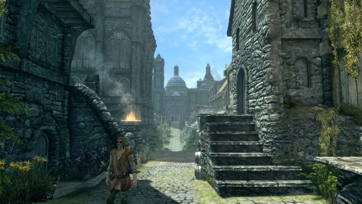 Image of Skyrim's city Solitude with the player on the main street. The Blue Palace building is visible in the background with an NPC approaching the player. It is a blue sky sunny day.