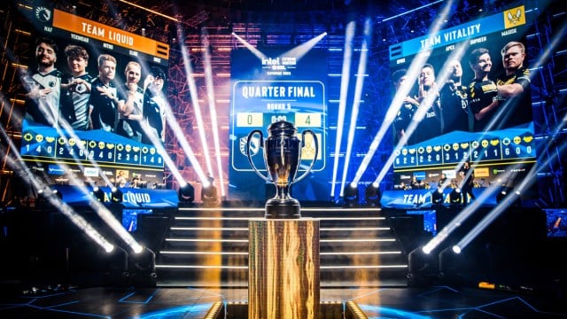 The IEM Katowice trophy on a plinth as two teams play Counter-Strike on-stage at the Spodek Arena.