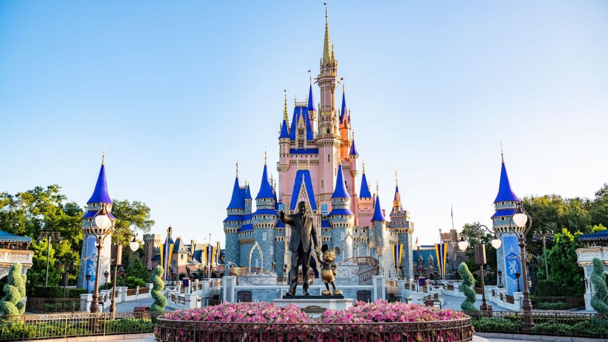 Image showcasing Cinderella's Castle at Disney World. There is a bright blue sky present on the day and a statue of Walt Disney standing next to Mickey Mouse is in the centre.