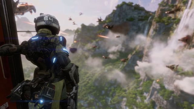 Image of a Pilot in Titanfall 2 standing at the edge of a chopper and looking out onto a forest mountainous biome in the background.