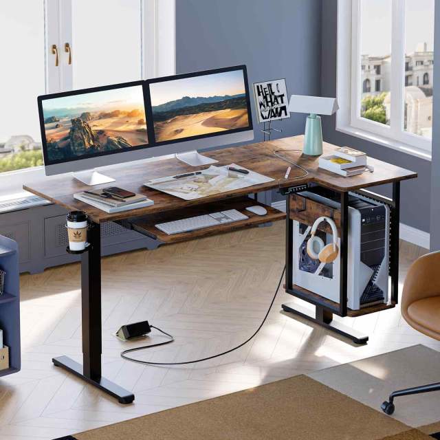Bestier L Shaped Electric Stand Up Desk with two monitors on top, a PC case, keyboard, and headset underneath it, in a gray room