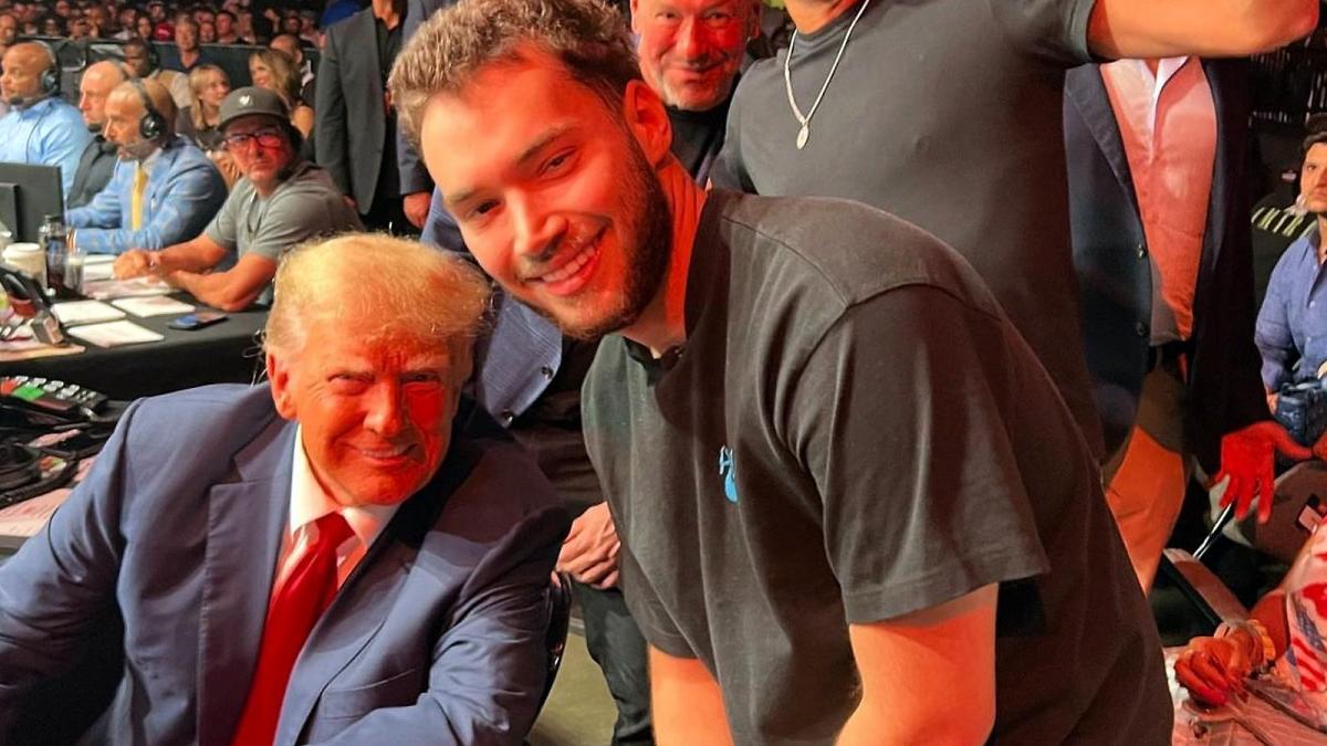Adin Ross stands next to former President Donald Trump at a UFC event while several people including Dana White look over in the crowd.
