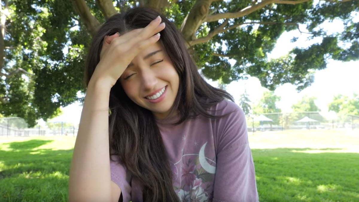 Pokimane sits outside in the grass with her hand over her head, looking embarrased.