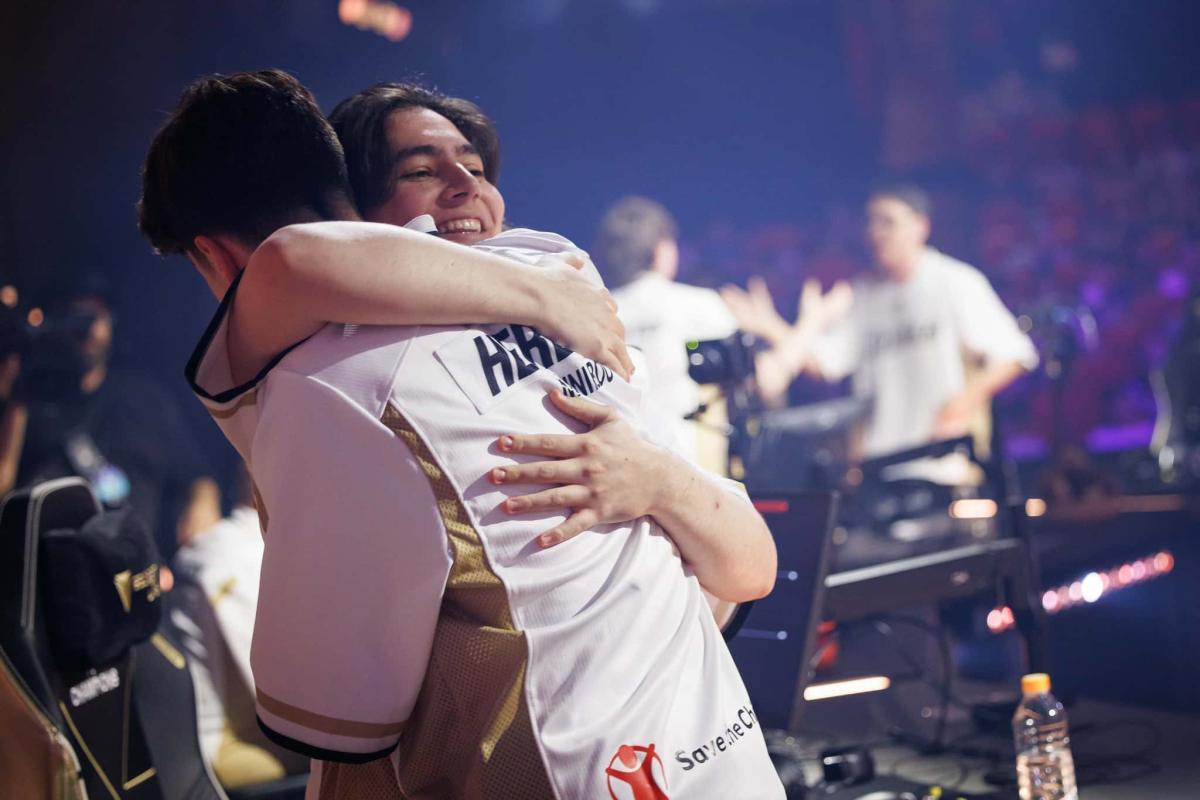 Two players of Team Heretics hug on stage after their win against FPX, behind them other players are celebrating; they're all wearing their new white jersey for the tournament