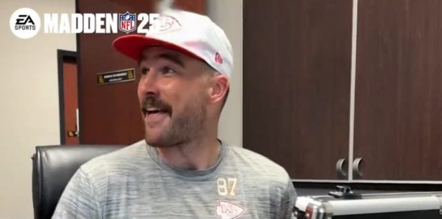 Travis Kelce smiles with a gray shirt and white hat, also sporting a mustache.