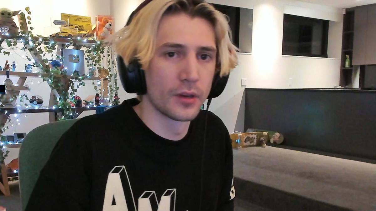 Felix "XQC" Lengyel sits in a nearly empty loungeroom on an office chair. There's several nick nacks and trinkets on a shelf behind him