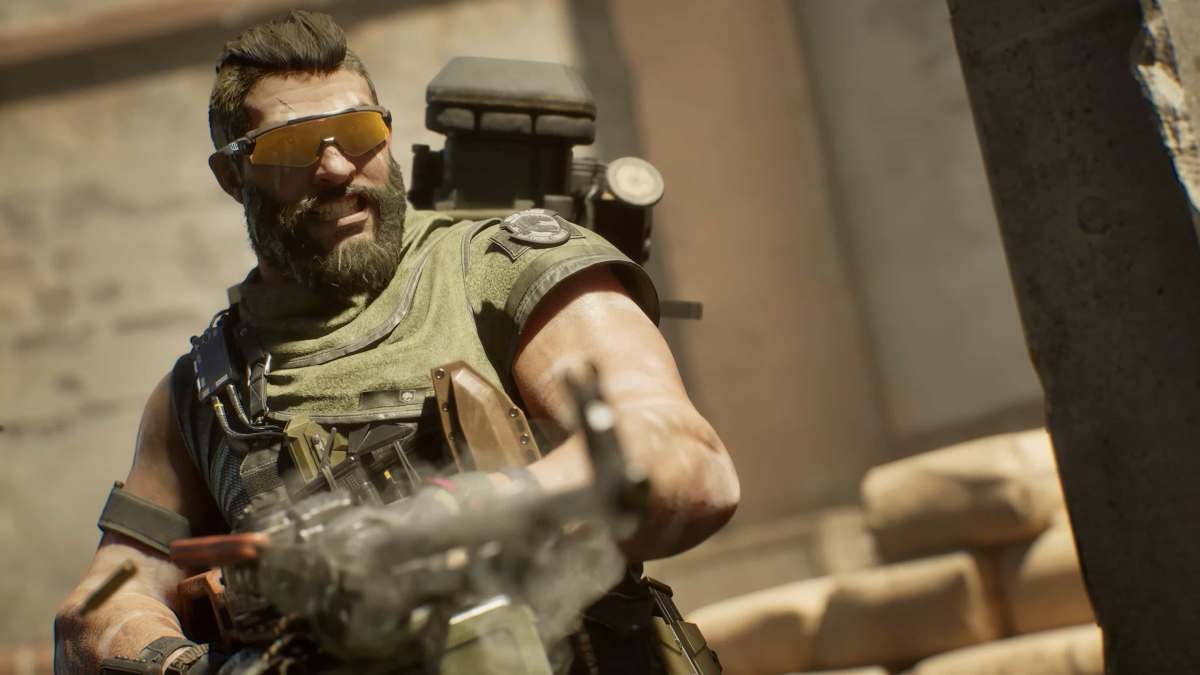 A bearded man in a bullet-proof vest and sunglasses shoots a large machine gun.