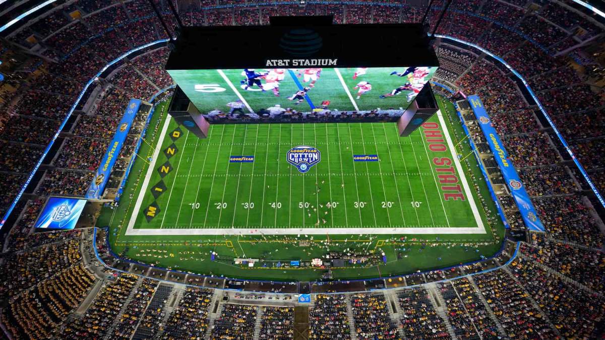 An overhead shot of a football field and large crowd watching a game, with a huge screen hanging over the field showing highlights.