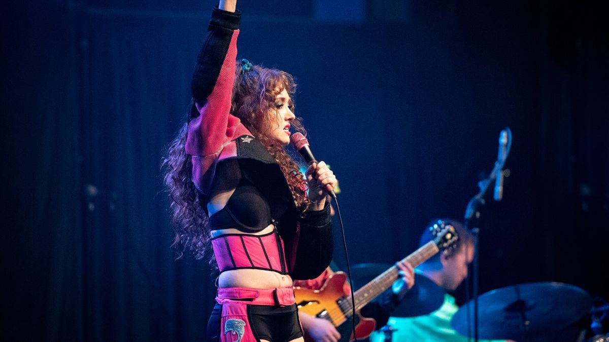 An image of Chappell Roan performing on a stage with a microphone and a pink and black outfit.