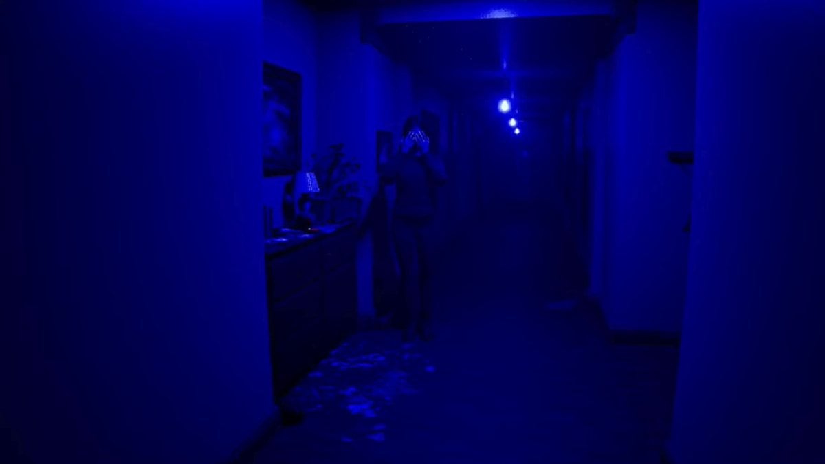 A blue lighting corridor with a woman standing off to the side, covering her face with her hands.