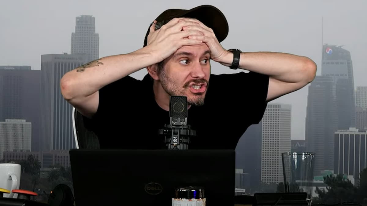 Ethan Klein wearing a hat and holding his hands to his head in anger and surprise with the skyline of NYC behind him.