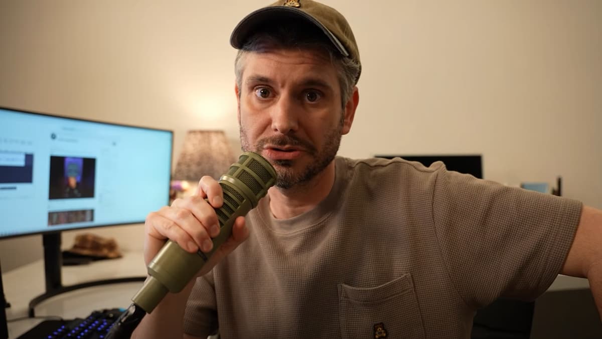 Ethan Klein holds a microphone with a computer screen in his background.